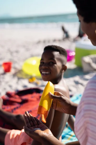 アフリカ系アメリカ人の男の子が笑顔で ビーチに座っている間に日焼け止めを持っている母親を見て コピースペース 変更されていない 一緒に ピクニック スキンケア 楽しみ — ストック写真