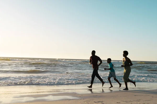 陽気アフリカ系アメリカ人の父と息子と母は日没時に空に対してビーチで実行しています コピースペース 変更されていない 一緒に 子供の頃 楽しみと夏 — ストック写真