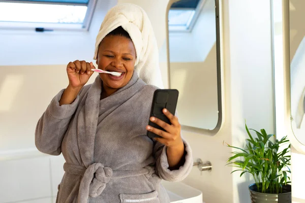 幸せプラスサイズアフリカ系アメリカ人女性は バスルームでローブを着て歯を磨き スマートフォンを使用しています セルフケア 健康と美しさ 身体的包容力 コミュニケーションとライフスタイル 変わらない — ストック写真