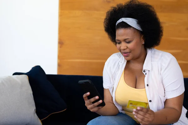 快乐加尺寸的非洲裔美国女人坐在沙发上 带着信用卡和智能手机在线购物 自由时间 家庭生活 身体包容和生活方式 — 图库照片