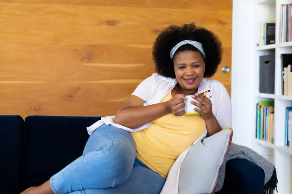 快乐加尺寸的非洲裔美国女人躺在沙发上 抱着杯子 微笑着 闲暇时间 家庭生活 身体包容和生活方式 — 图库照片
