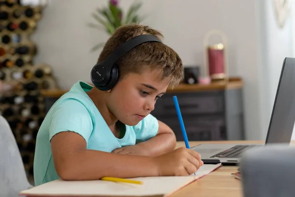 自宅のテーブルの上にノートパソコンで宿題を書いてヘッドフォンを着て白人の少年 コピースペース 変更されていない 子供時代 無線技術 学生およびEラーニングの概念 — ストック写真