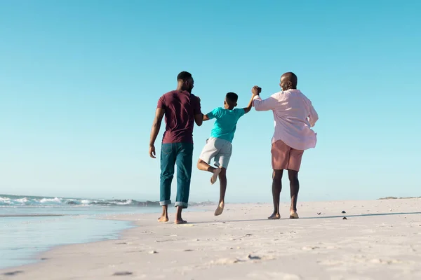 アフリカ系アメリカ人の父親と祖父が少年の手を握って海岸で彼を拾い上げる様子 コピースペース 変更されていない 多世代の家族 一緒に 楽しむ — ストック写真