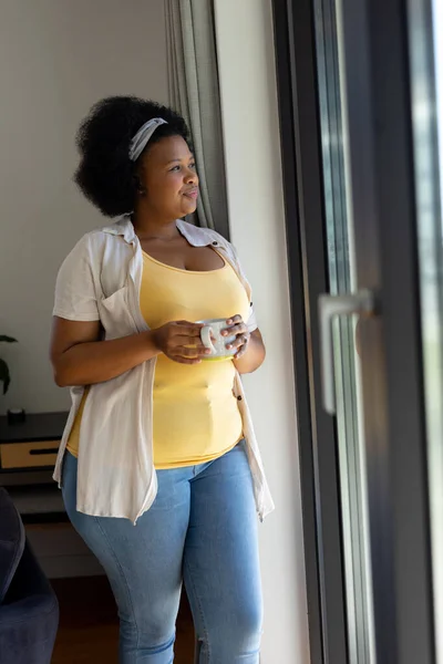 有思想和体形的非洲裔美国女人举着杯子向窗外望去 家庭生活 身体包容和生活方式 — 图库照片