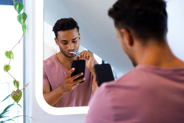 Biracial man in mirror, brushing teeth and using smartphone in bathroom. Lifestyle, self care, communication and domestic life, unaltered.