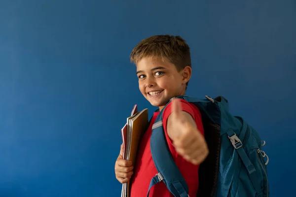 青い背景の上に親指を示す本や袋を持つ白人の笑顔の少年 スペースをコピー 変更されず ライフスタイル 子供時代 学校の少年 正の記号と幸せなコンセプト — ストック写真
