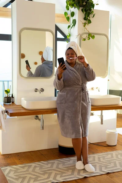 幸せプラスサイズアフリカ系アメリカ人女性は バスルームでローブを着て歯を磨き スマートフォンを使用しています セルフケア 健康と美しさ 身体的包容力 コミュニケーションとライフスタイル 変わらない — ストック写真