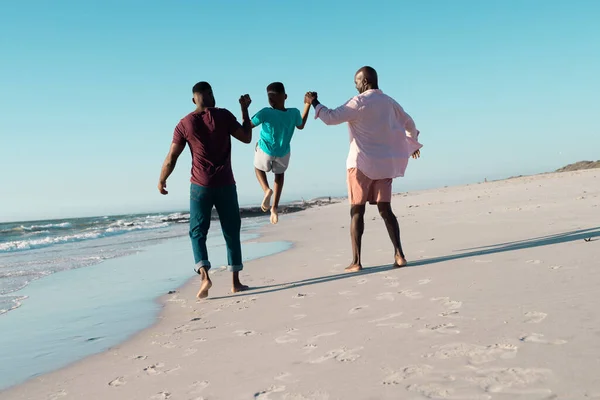 stock image Rear view of african american father and grandfather holding boy's hands and picking him up at beach. Copy space, unaltered, multigeneration family, together, child, nature, vacation, enjoy, summer.