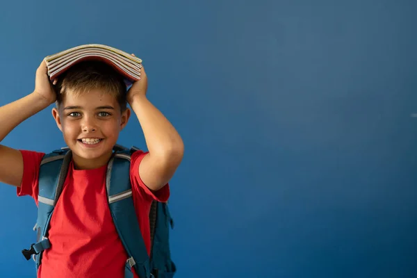 青い背景の上に立っている間 頭の上に本を運ぶ笑顔の白人少年の肖像画 コピースペース 変更されていない ライフスタイル 子供時代 学校の少年 学生と幸せなコンセプト — ストック写真