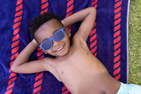サングラスをかけタオルに寝そべって笑顔を見せるアフリカ系アメリカ人の高いアングル 変更されていない ライフスタイル 子供時代 頭の後ろの手 リラックスして楽しむコンセプト — ストック写真