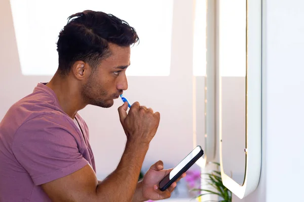 Biracial Man Tittar Spegeln Borsta Tänderna Och Använda Smartphone Badrummet — Stockfoto