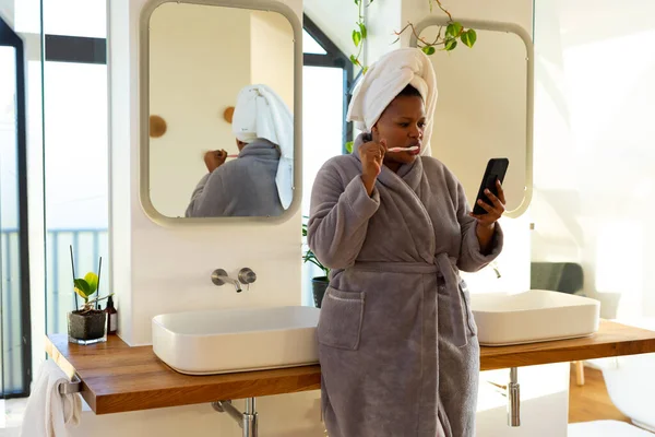 플러스 사이즈 아프리카 미국인 가운입고 양치하고 화장실에서 스마트폰 자기보살핌 과아름다움 — 스톡 사진