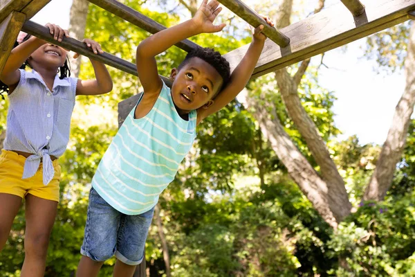 stock image Low angle view of african american playful siblings hanging on monkeys bars at playground. Unaltered, family, togetherness, childhood, playing, enjoyment and weekend concept.