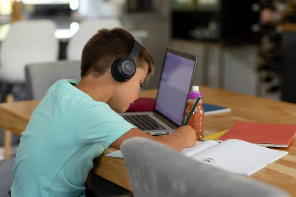 自宅のテーブルの上にノートパソコンで宿題を書いてヘッドフォンを着て白人少年 コピースペース 変更されていない 子供時代 無線技術 オンライン Eラーニングの概念 — ストック写真