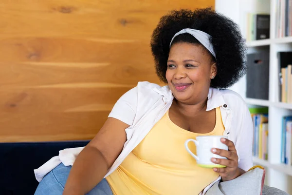 ハッピープラスサイズアフリカ系アメリカ人女性ソファ保持マグカップでリラックス リラクゼーション 自由な時間 国内生活 身体的包容力とライフスタイル 変更されない — ストック写真