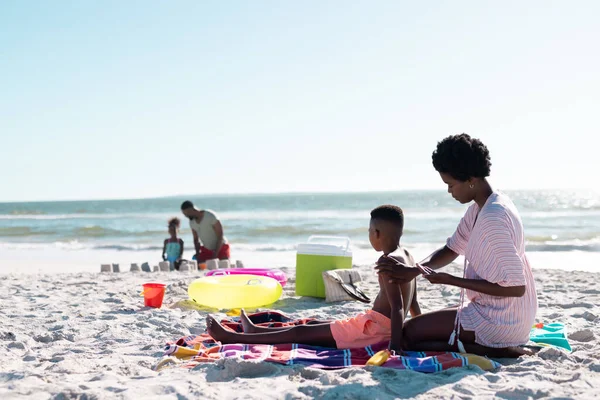 アフリカ系アメリカ人の女性は 息子の体に日焼け止めを塗る一方で 空に対してビーチに座っている コピースペース 変更されていない 一緒に ピクニック スキンケア 楽しみ — ストック写真