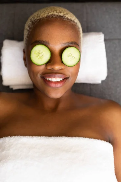 快乐的非洲裔美国女人躺在毛巾上 眼睛上长着黄瓜片 美感和生活方式概念 — 图库照片
