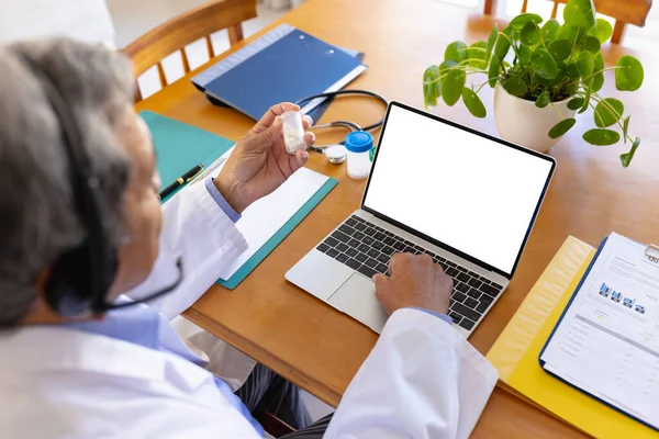 Starszy Dwurasowy Lekarz Trzymający Butelkę Pigułek Pomocą Laptopa Zestawu Słuchawkowego — Zdjęcie stockowe
