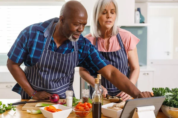 stock image Happy senior diverse couple wearing aprons and using tablet in kitchen. Spending quality time at home and retirement concept.