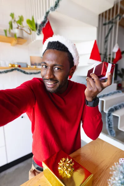 サンタの帽子をかぶっている幸せなアフリカ系アメリカ人の男性は 現在を保持し ビデオ通話をしています クリスマスだけで家で質の高い時間を過ごす — ストック写真