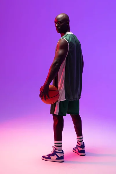 Image of portrait of african american basketball player with basketball on neon purple background. Sports and competition concept.