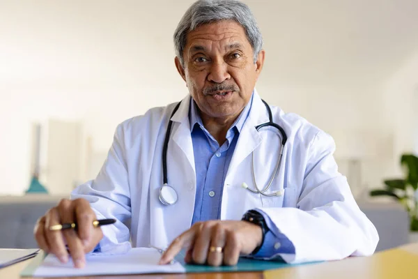 Starszy Dwurasowy Lekarz Siedzący Przy Biurku Rozmawiający Podczas Konsultacji Wideo — Zdjęcie stockowe