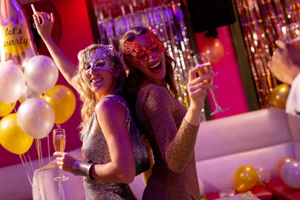 仮面をかぶった2人の幸せな白人女性の友人が ナイトクラブのパーティーでシャンパンを飲みながら踊ります 楽しさ お祝い パーティーのコンセプト — ストック写真