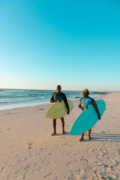 海と青い空に対して砂浜を歩くサーフボードを持つアフリカ系アメリカ人のシニアカップル コピースペース ウォータースポーツ レクリエーション 変更なし 一緒に お楽しみください — ストック写真