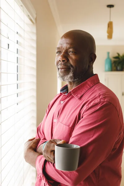 コーヒーの笑顔で窓のそばに立つ思慮深いアフリカ系アメリカ人の先輩の垂直イメージ 包摂性 国内生活の概念 — ストック写真