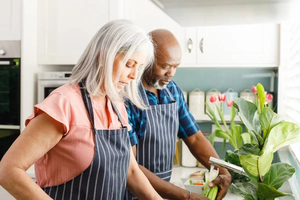 台所でエプロンと料理を身に着けている幸せな高齢者の多様なカップル 家庭や退職のコンセプトで質の高い時間を過ごす — ストック写真