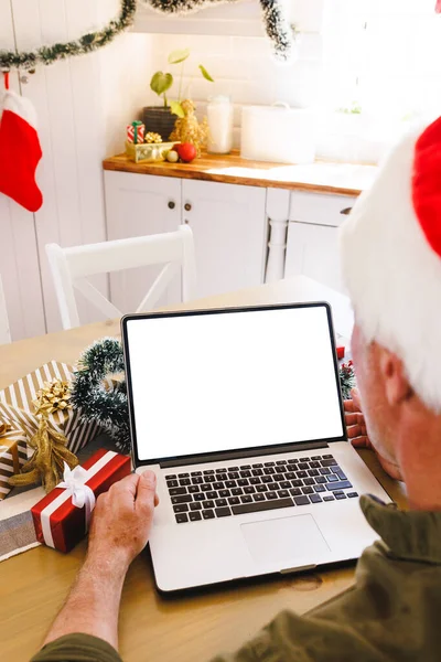 サンタの帽子をかぶった白人男性がキッチンのテーブルに座り コピースペース付きのノートパソコンを使用しています クリスマスだけで家で質の高い時間を過ごす — ストック写真
