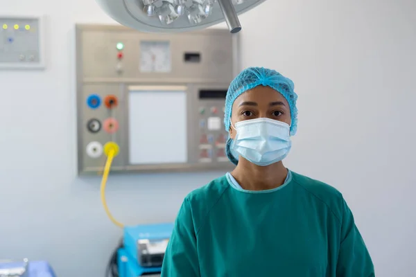 Portrait of biracial female healthcare worker in surgical cap, mask and gown in operating theatre. Hospital, medical and healthcare services.