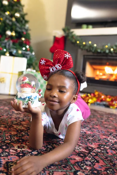 非洲裔美国女孩拿着圣诞雪球坐在隔壁壁炉的垂直图片 圣诞节 童年和庆祝概念 — 图库照片