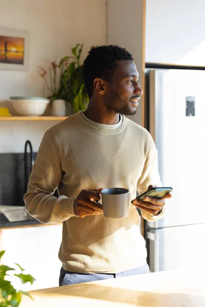 キッチンに立ち コーヒーを飲み スマートフォンを使う幸せなアフリカ系アメリカ人男性 自宅だけで質の高い時間を過ごす — ストック写真