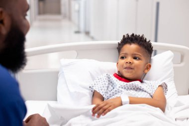 African american male doctor talking to boy patient in hospital bed in corridor with copy space. Hospital, medical and healthcare services. clipart