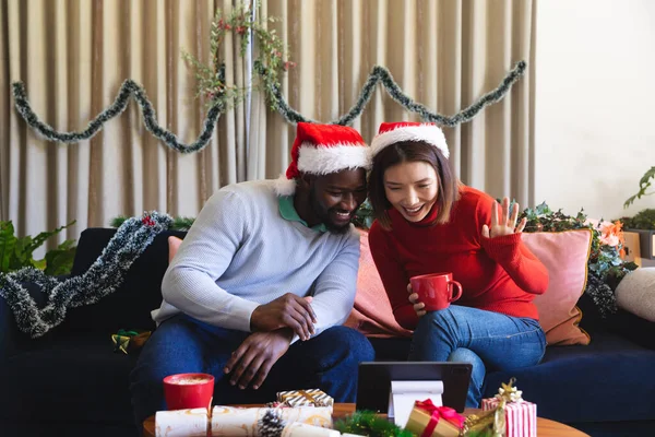 ビデオ通話にタブレットを使用して サンタクラスの帽子を身に着けている幸せな多様なカップル 一緒にクリスマスに質の高い時間を過ごす — ストック写真