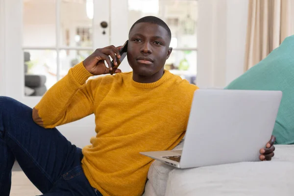 アフリカ系アメリカ人の男性がノートパソコンを使い 自宅のソファで床に座ってスマホで話しています 包括性 国内生活 遠隔作業 — ストック写真