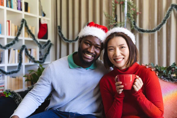 カメラと笑顔を見て サンタクラスの帽子をかぶって幸せな多様なカップルの肖像画 一緒にクリスマスに質の高い時間を過ごす — ストック写真