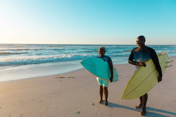アフリカ系アメリカ人のシニアカップルは 砂浜を歩いて空に向かってサーフボードを持っています コピースペース ウォータースポーツ レクリエーション 変更なし 一緒に 楽しみ — ストック写真
