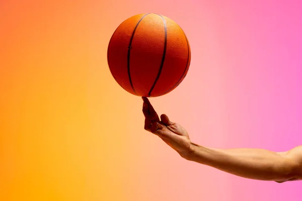Image of hand of biracial basketball player spinning basketball on pink to orange background. Sports and competition concept.