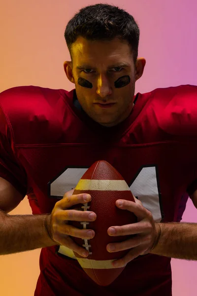 Caucasian male american football player holding ball with neon yellow and purple lighting. Sport, movement, training and active lifestyle concept.
