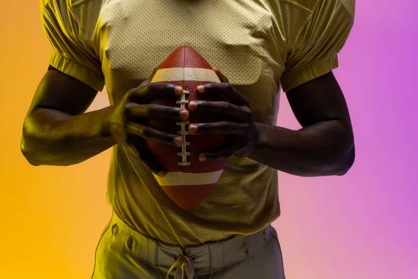 Midsection of african american male american football player with neon purple and yellow lighting. Sport, movement, training and active lifestyle concept.