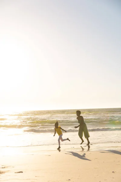 日没時に澄んだ空の下でビーチを走る遊び心のあるアフリカ系アメリカ人の母親と娘 コピースペース 変更されていない 一緒に 楽しみ 子供時代と自然コンセプト — ストック写真