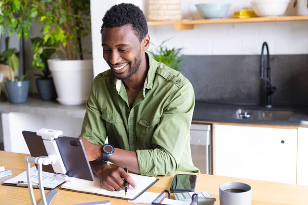 stock image Happy african american man standing at table in kitchen, using tablet. Spending quality time at home alone.