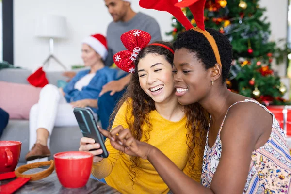 Image of happy diverse female friends using smartphone, celebrating christmas with diverse friends. Christmas, celebration, tradition, friendship, inclusivity and lifestyle concept.