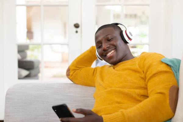 Sonriente Hombre Afroamericano Casa Sentado Sofá Con Auriculares Mirando Teléfono —  Fotos de Stock