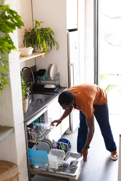 キッチンに立って食器洗い機をアンロードする幸せなアフリカ系アメリカ人男性 自宅だけで質の高い時間を過ごす — ストック写真