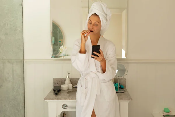 幸せな白人女性は バスルームでローブを着て歯を磨き スマートフォンを使用しています 質の高い時間を家だけで過ごす — ストック写真