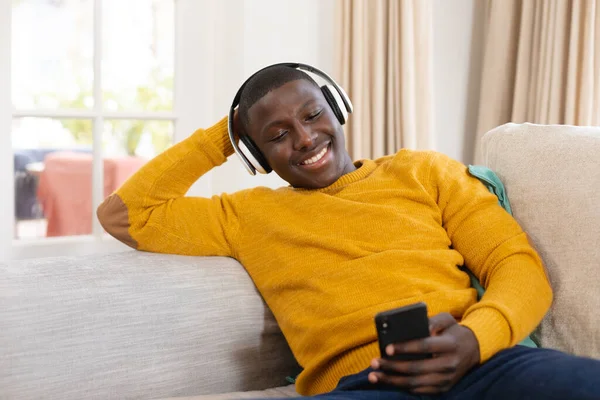 Gülümseyen Afro Amerikan Adam Evde Koltukta Oturmuş Kulaklık Takıyor Akıllı — Stok fotoğraf