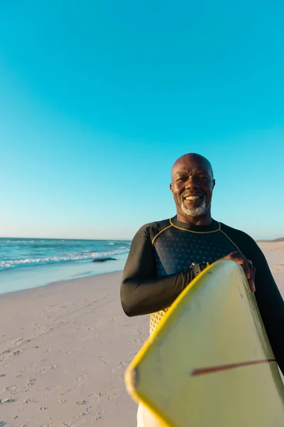 秃头的非洲裔美国老人背着冲浪板站在海滩上 面对蓝蓝的天空 复制空间 水上运动 — 图库照片